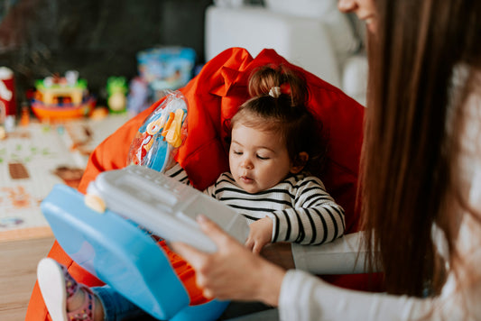 Małe dziecko siedzi na czerwonej pufie i bawi się zabawkowym laptopem, a obok dorosła osoba pomaga mu w zabawie.