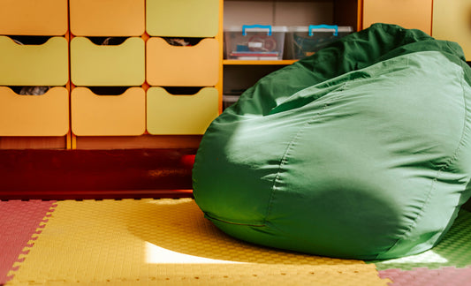 Zielona pufa typu worek w pokoju dziecięcym na piankowej macie, w tle żółte szafki z szufladami.