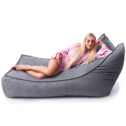 Kobieta leżącą bokiem na dużej, szarej pufie leżance Horizont, wykonanej z tkaniny imitującej len.