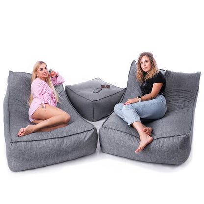 Dwie kobiety siedzące na fotelach HORIZONT z szarej tkaniny len z pufą-stolikiem pośrodku.