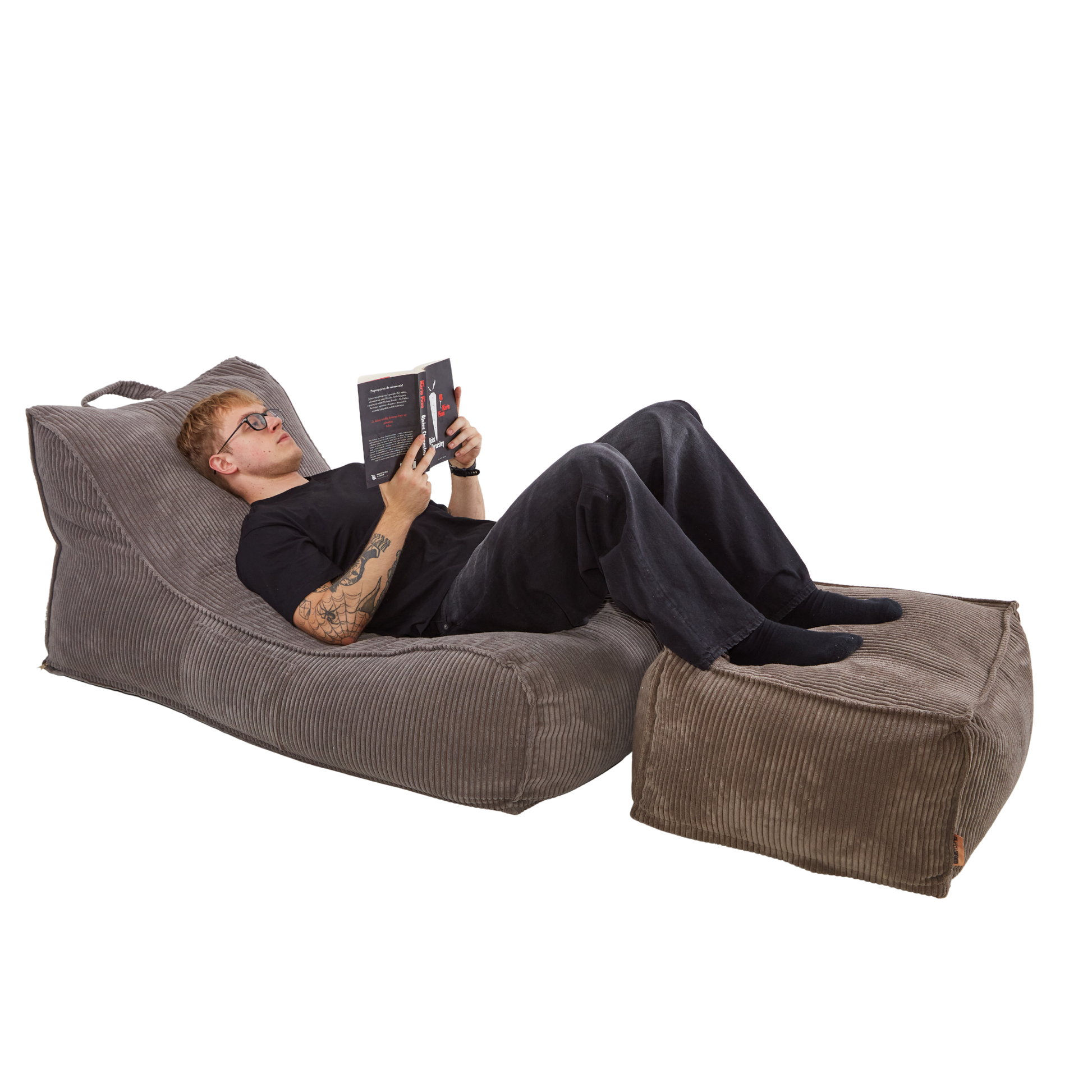 Mężczyzna czytający książkę na sztruksowej leżance Canvas z nogami opartymi o pufę. Całość wykonana jest z tkaniny sztruksowej w kolorze jasnego brązu. 