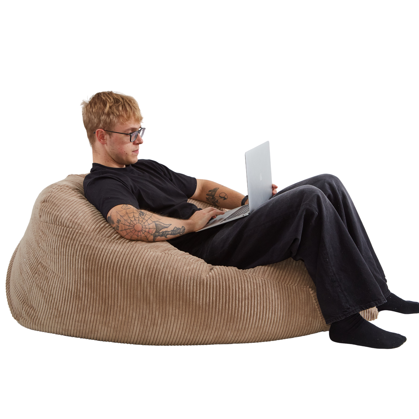 Mężczyzna pracujący na laptopie, siedząc na dużej sztruksowej pufie-poduszce w kolorze toffi. 