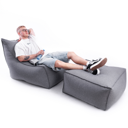 Mężczyzna odpoczywający na ogrodowinym, wodoodpornym fotelu LENO z nogami wyłożonymi na dużej pufie do komplety. Zestaw na zdjęciu jest w kolorze szarym, z tkaniny imitującej len.