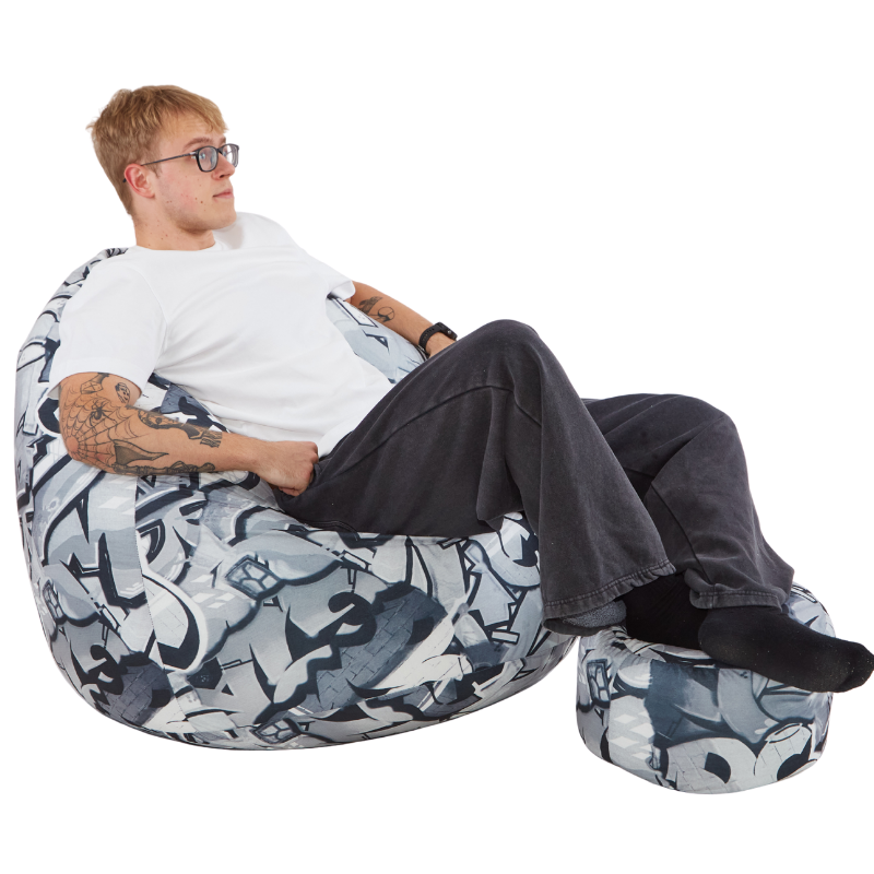 Mężczyzna siedzący na fotelu typu worek sako we wzór z szarym graffiti, z nogami położnymi na pufie w ten sam wzór. 